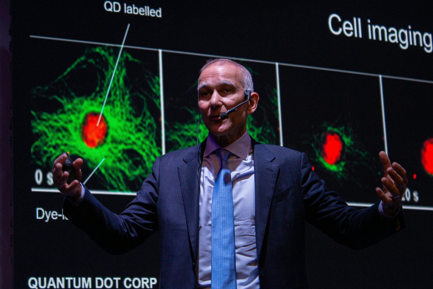 Presentatore in giacca e cravatta che spiega grafici scientifici su uno schermo.